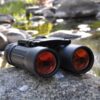 Black compact binoculars with ruby red lenses resting on a rock beside a stream